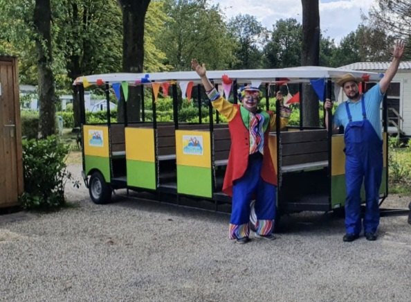 Het treintje van de camping met vlaggetjes, met twee vrolijke medewerkers ervoor